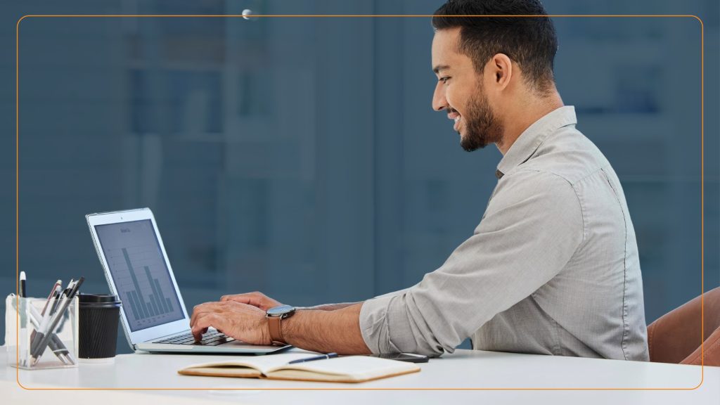 A smiling man types on a laptop at a bright office desk with a graph visible on his screen. The setup suggests business productivity and tech engagement, representing the usability and efficiency of the best TCPA compliance software.